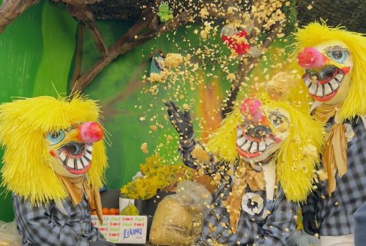 Basler Fasnacht 2025: Die „drey scheenschte Dääg“ Drei Fasnachtsteilnehmer mit grossen, bunten Masken, gelben Haaren und karierten Kostümen werfen Konfetti in die Luft, typisch für die Basler Fasnacht.