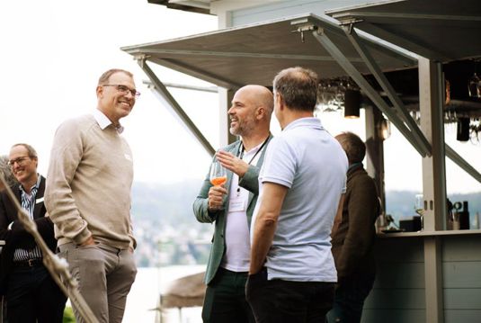 Drei Männer im Gespräch bei lockerer Atmosphäre am Coopers Summer Apéro, mit Blick auf den Zürichsee im Hintergrund.
