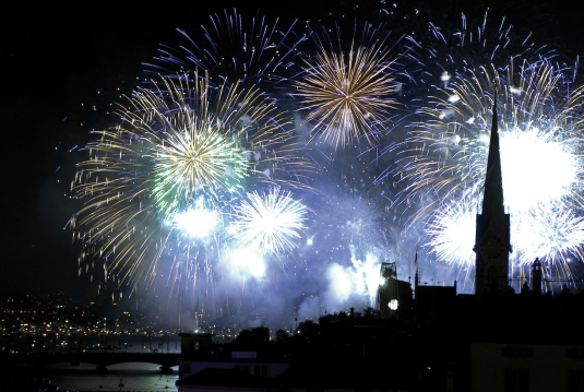 Fireworks over Lake Zurich at night with the illuminated old town and a prominent church tower in the foreground.