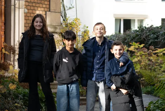 Vier Kinder stehen draussen nebeneinander und lächeln in die Kamera. Sie tragen Herbst- oder Winterkleidung und stehen vor einem Gebäude mit Pflanzen im Hintergrund.