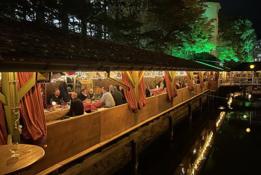 Abendliches Fondue in einer warm beleuchteten Holzhütte direkt am Wasser, Gäste sitzen in kleinen Gruppen gemütlich zusammen.