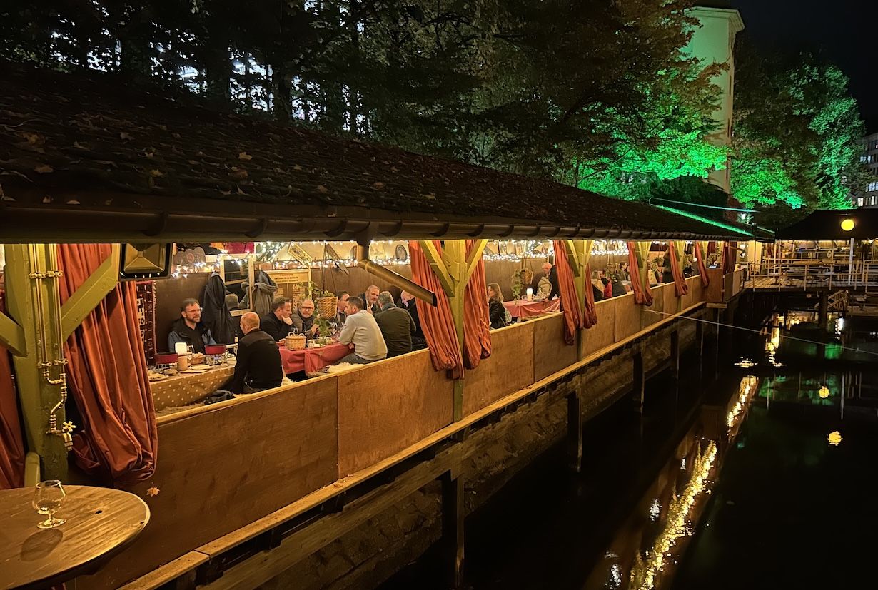 Abendliches Fondue in einer warm beleuchteten Holzhütte direkt am Wasser, Gäste sitzen in kleinen Gruppen gemütlich zusammen.