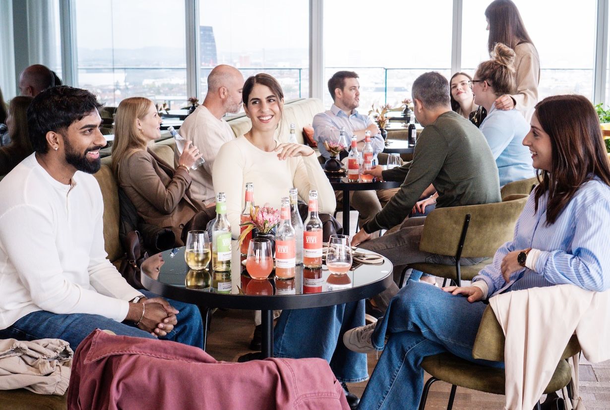 Coopers Team sitzt in gemütlicher Lounge-Runde an kleinen Tischen vor grossen Fenstern, mit Getränken und angeregten Gesprächen m B1 Rooftop Bistro in Basel.
